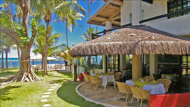 Un área de comedor al aire libre con sombrillas de paja, mesas y sillas, con vista a una playa y palmeras.