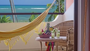 Un balcón con vistas al océano, una silla de mimbre, una mesita con bebidas y fruta, y una hamaca.