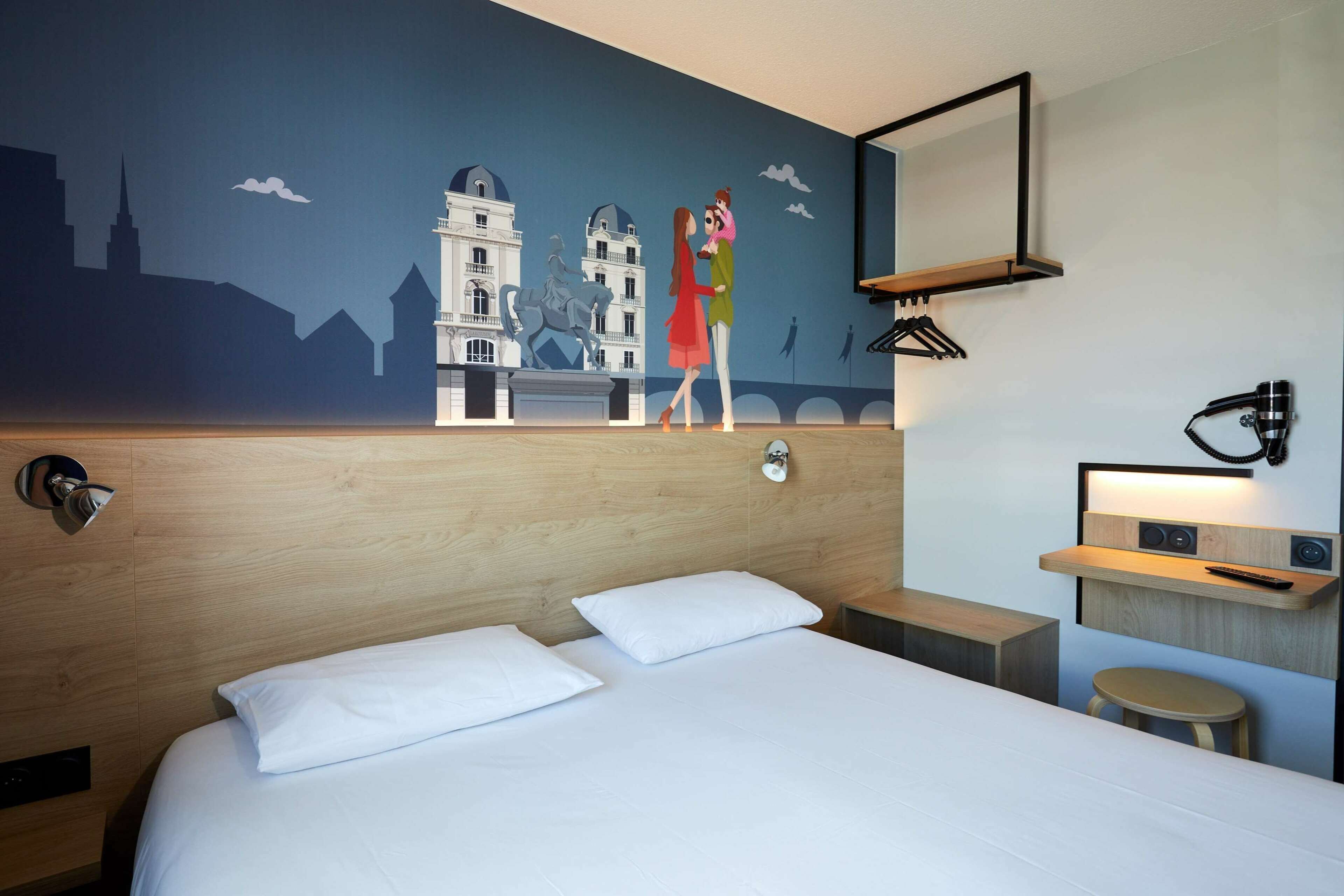 Una habitación de hotel con cama, mesita de noche, estante en la pared, espejo y un mural que muestra un paisaje urbano.