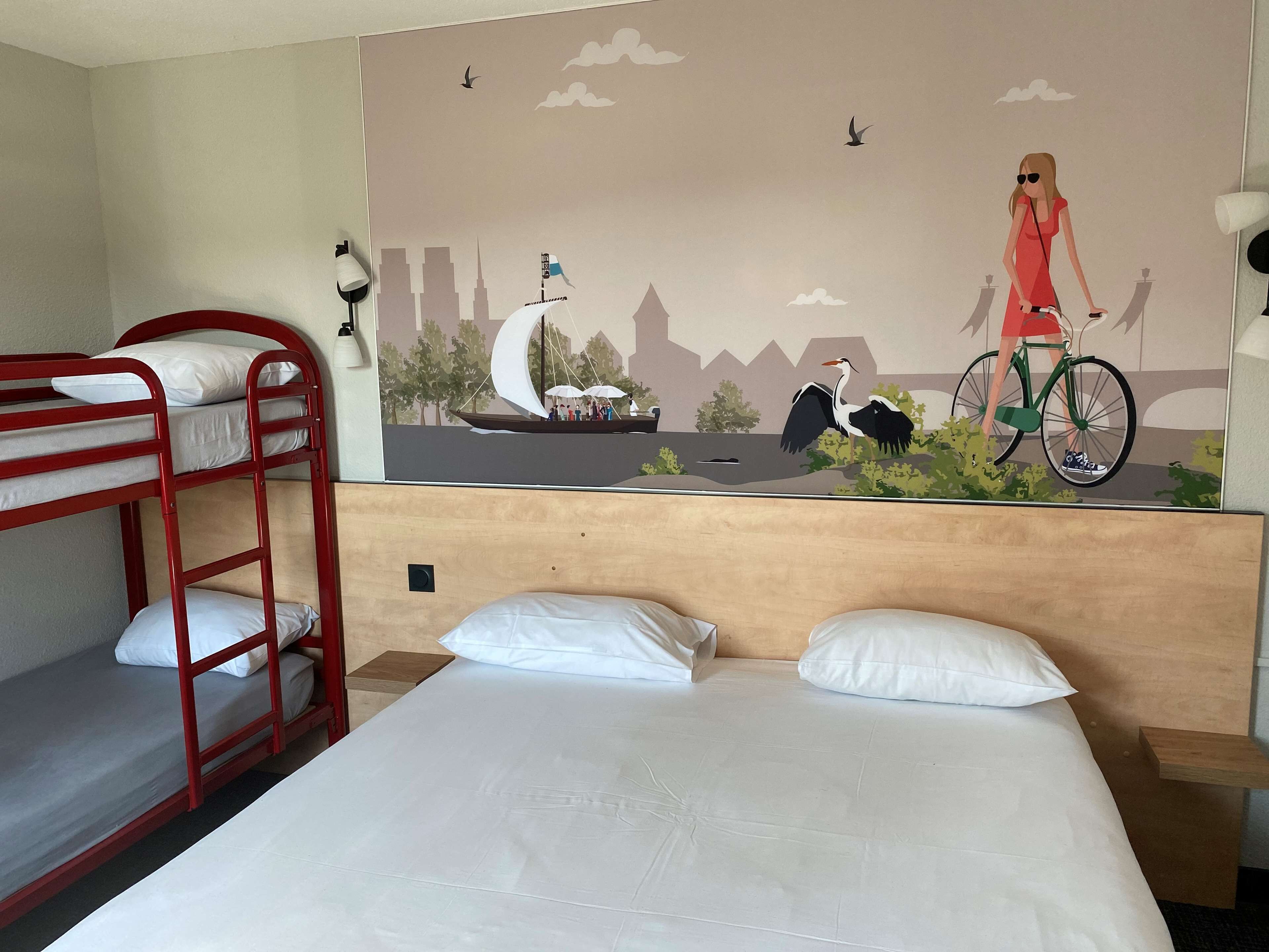 A hotel room with a bunk bed, a single bed, a mural depicting a cityscape with a cyclist, and a nightstand with a lamp.