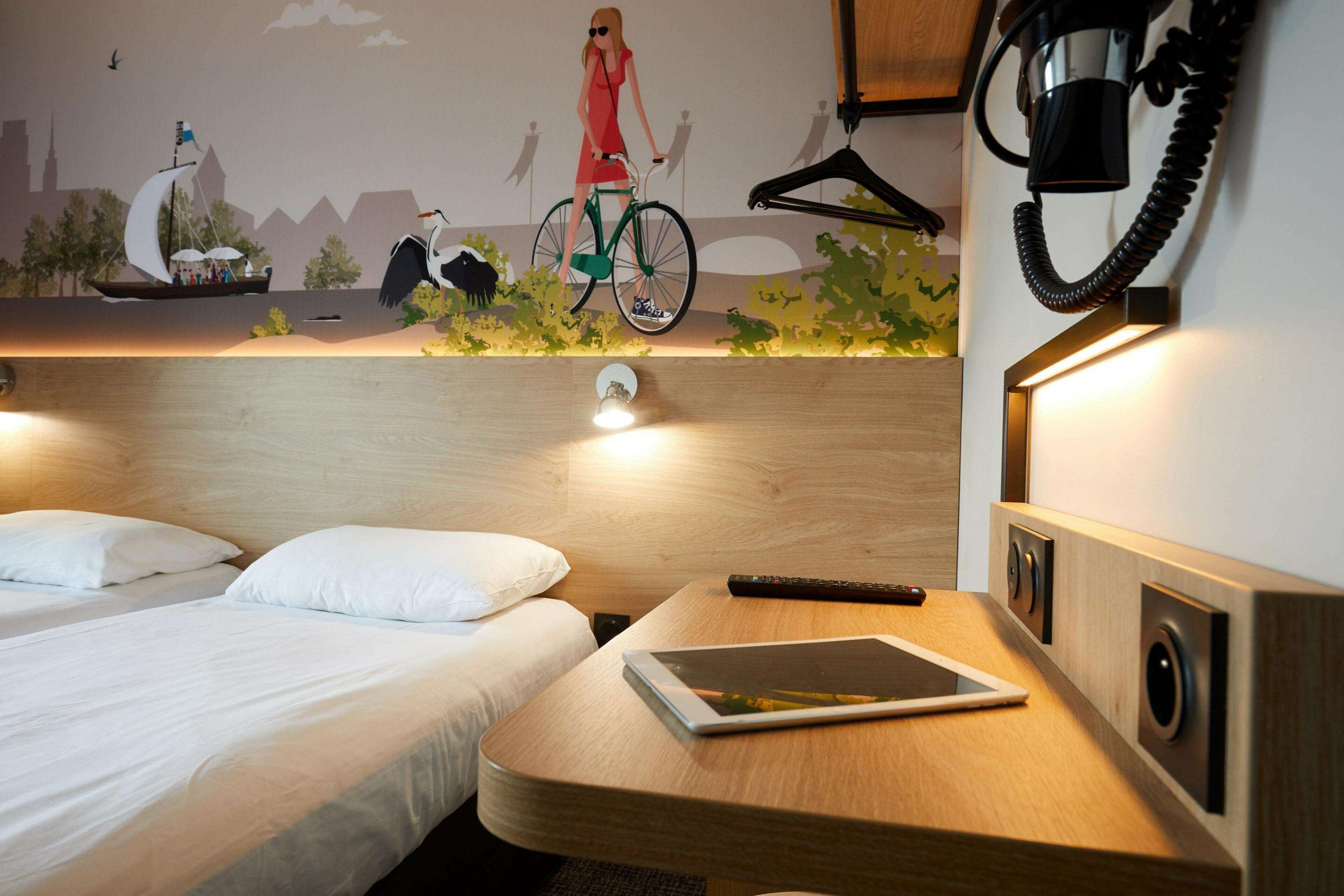 Camera d'albergo con due letti, un comodino con un tablet, un telefono a muro e un murale raffigurante una donna in bicicletta.