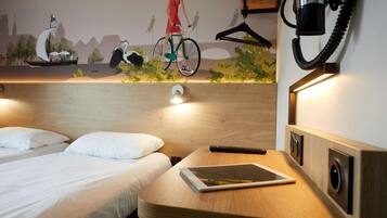 Camera d'albergo con due letti, un comodino con un tablet, un telefono a muro e un murale raffigurante una donna in bicicletta.
