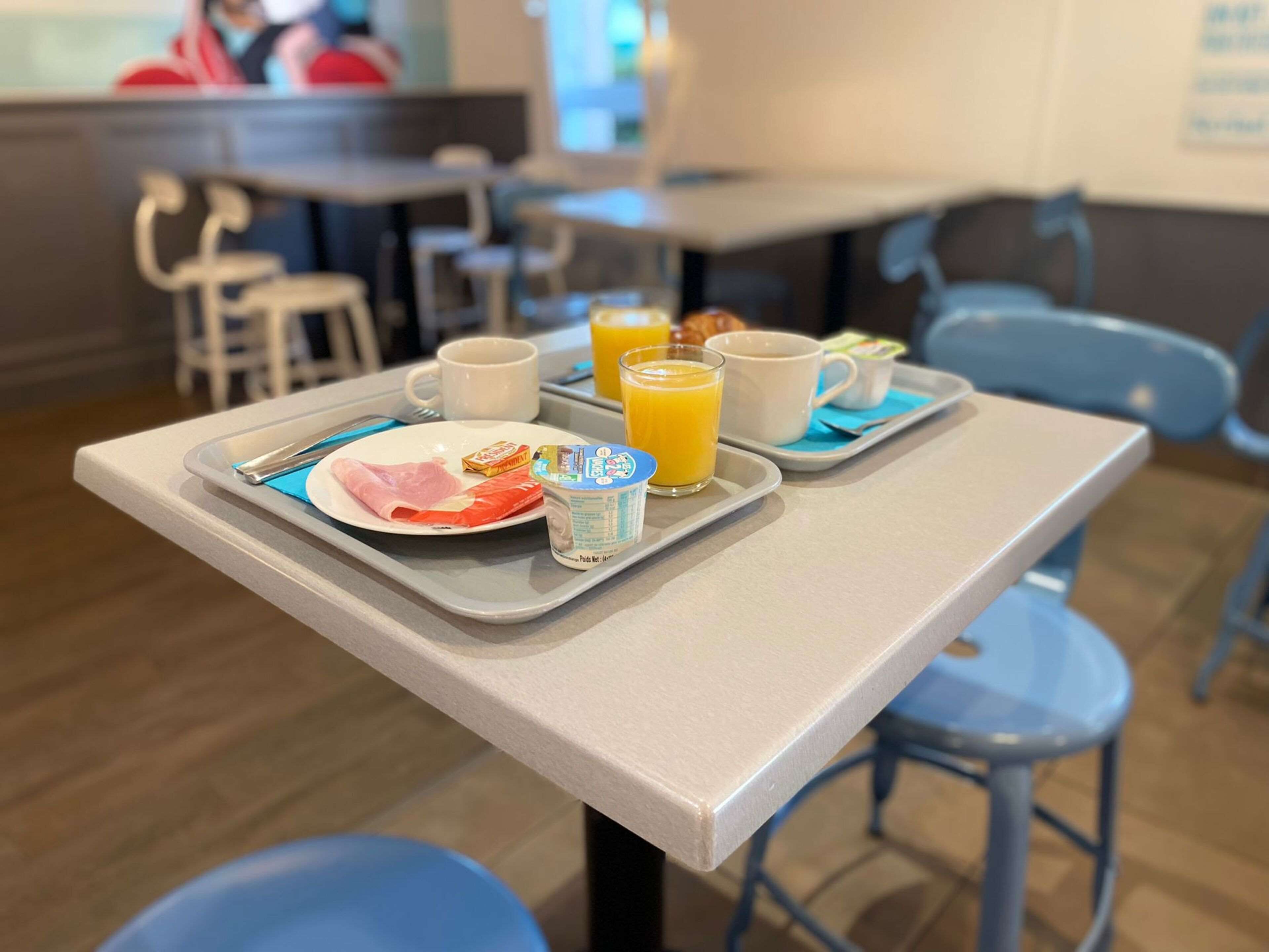 Una bandeja de desayuno con un plato de comida, un vaso de zumo de naranja y un vaso de yogur sobre una mesa en un comedor con sillas azules y una encimera al fondo.