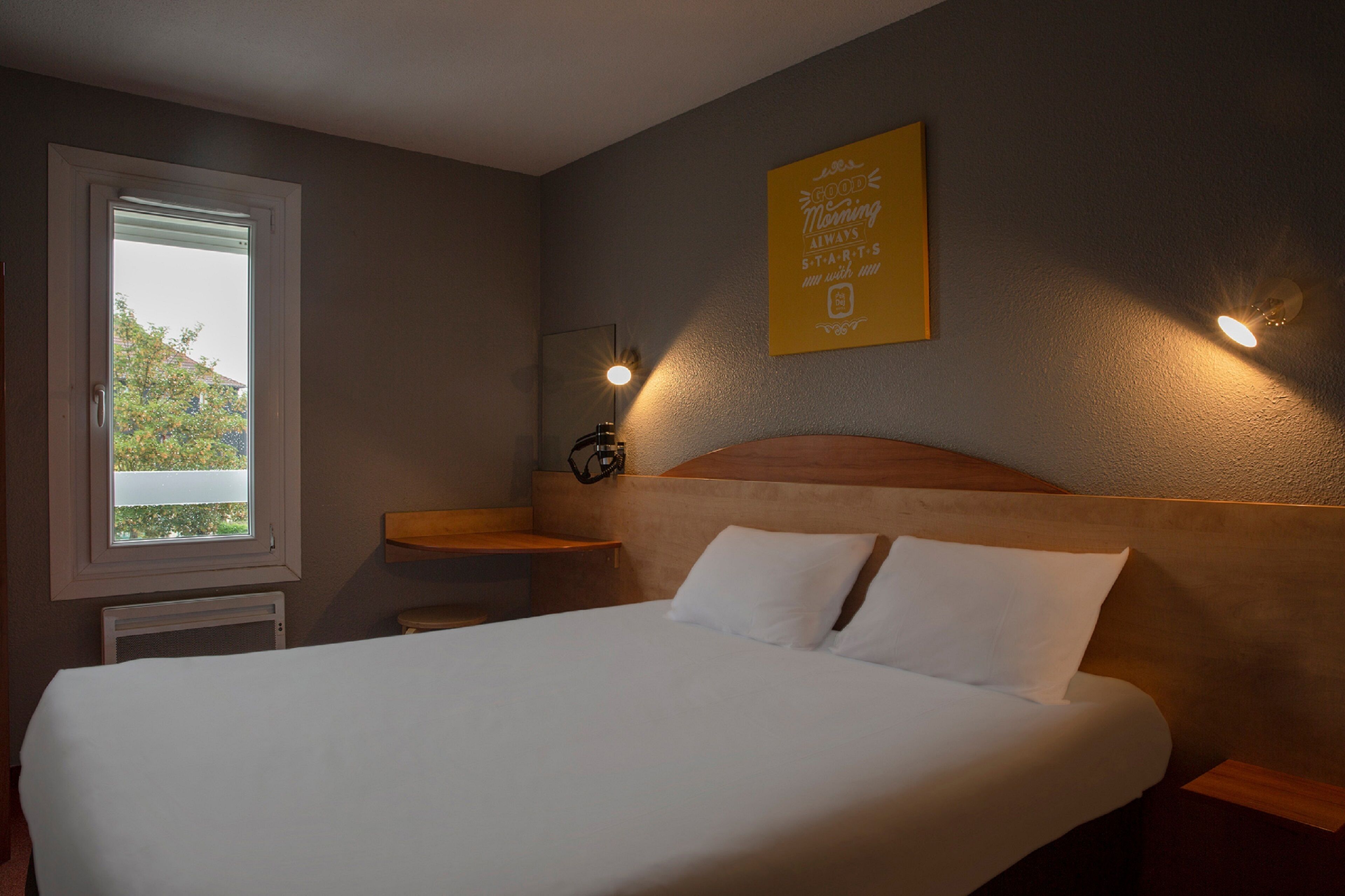 A neatly made bed in a hotel room with a window, a bedside table, and a wall-mounted light fixture.