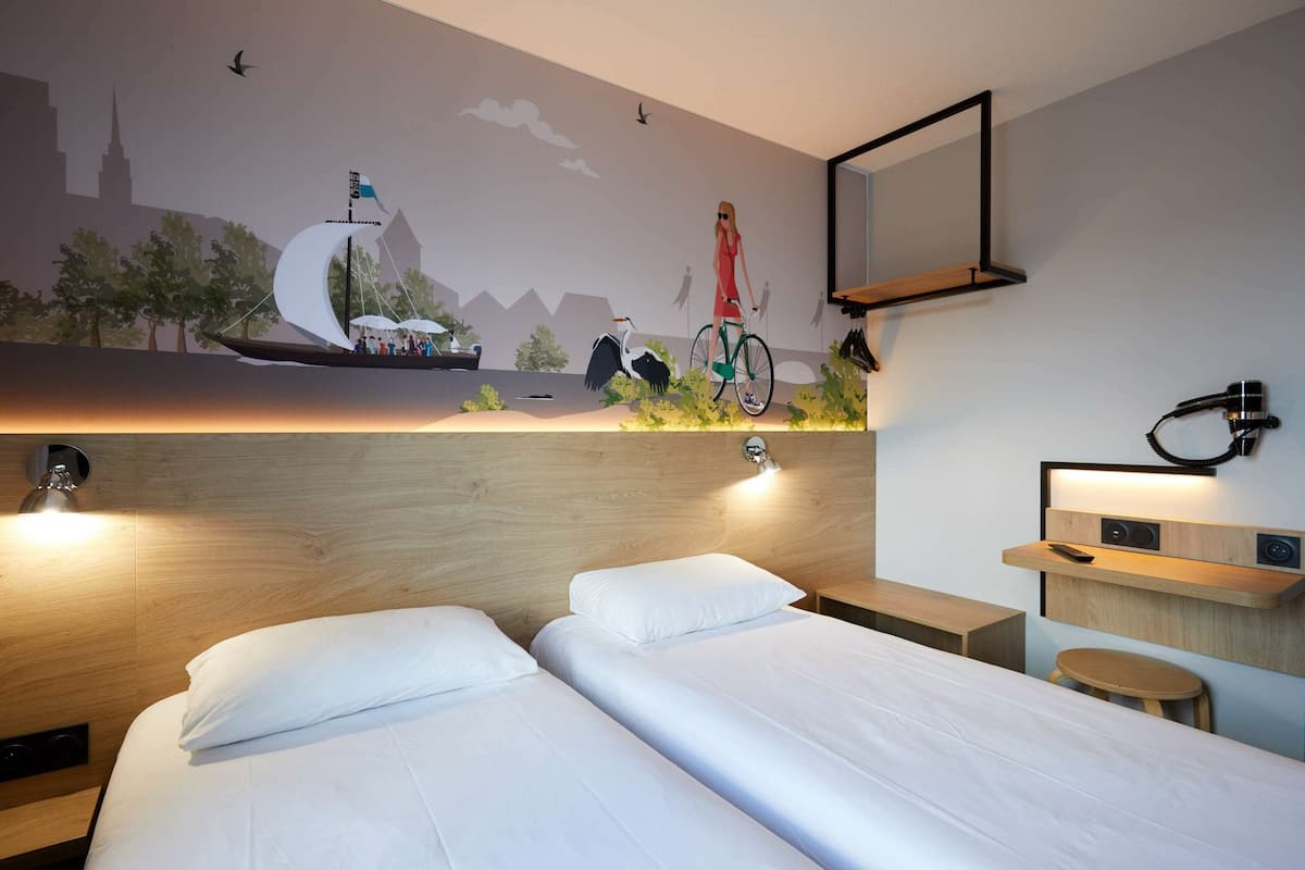 Ein modernes Hotelzimmer mit einem Holzkopfteil, zwei Betten, einem kleinen Schreibtisch und einem Wandgemälde, das eine Stadtansicht mit Segelboot und Radfahrer zeigt.