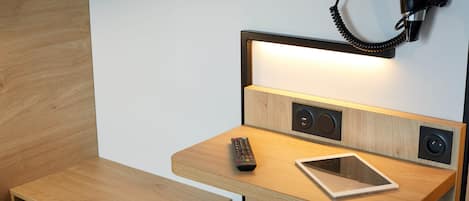 Una mesita de noche de madera con un control remoto, una tableta y una lámpara de pared.
