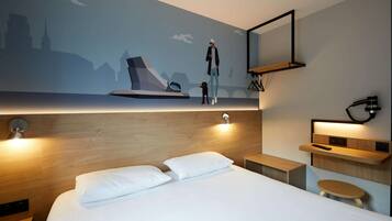 Quarto de hotel moderno com uma cama grande, cabeceira de madeira, televisão fixada na parede e uma mesinha.