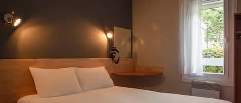 A hotel room with a large bed, a bedside table, a nightstand, a wall-mounted lamp, a window with a white curtain, and a view of greenery outside.