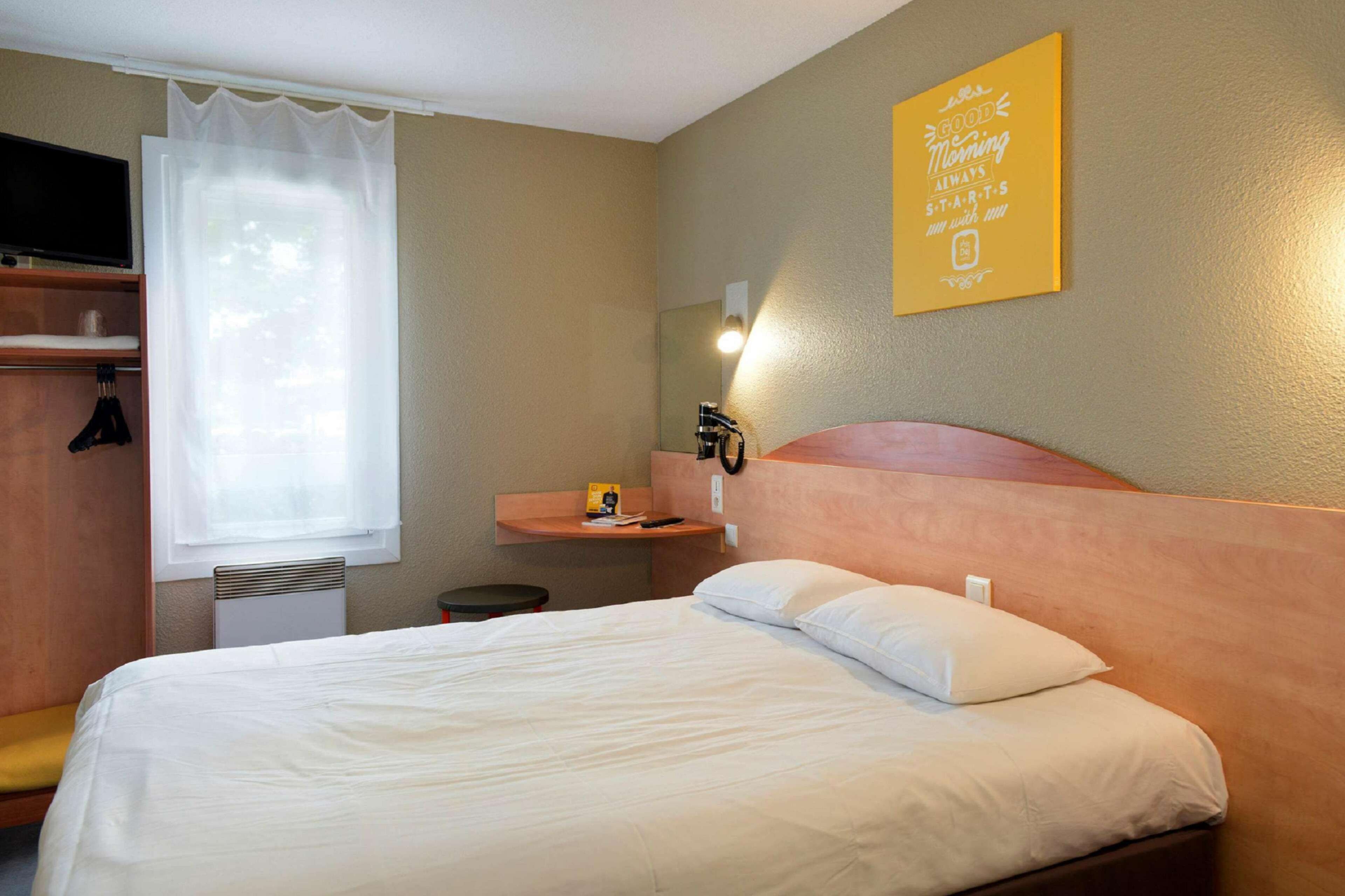 A hotel room with a bed, a bedside table, a mirror, a window with curtains, and a motivational poster on the wall.