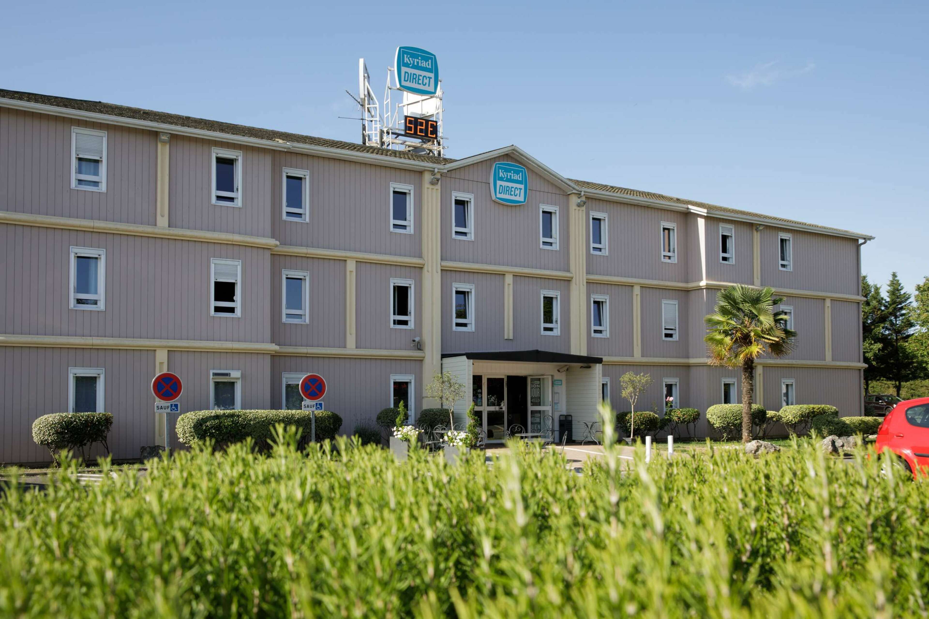 Un hotel de varios pisos con un letrero que indica 'Kyriad Direct' y una pantalla digital que muestra '520'. No se observan ventanas en la fachada, y el hotel tiene un techo plano.