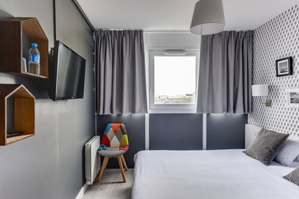 A modern hotel room with a large bed, a TV mounted on the wall, a chair with a colorful cushion, and a window with curtains.