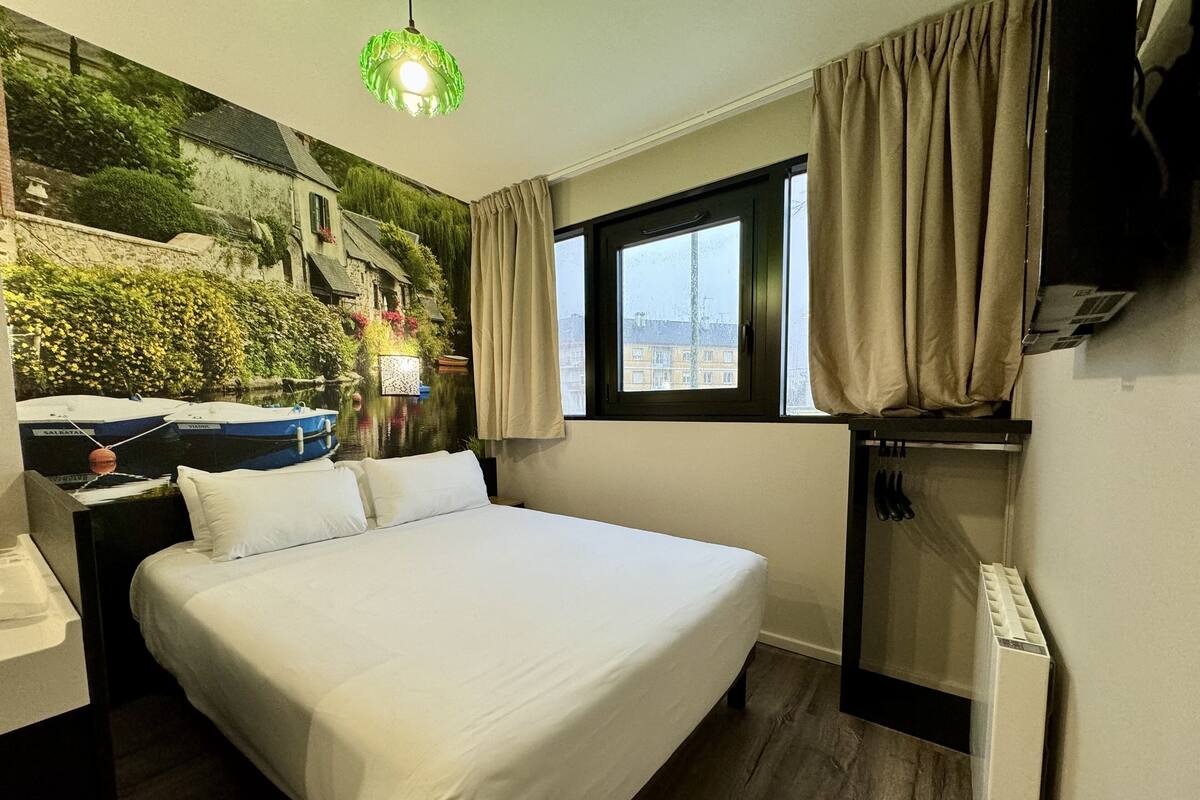 A hotel room with a bed, a desk, a chair, a TV, a window with curtains, and a mural of boats on the wall.