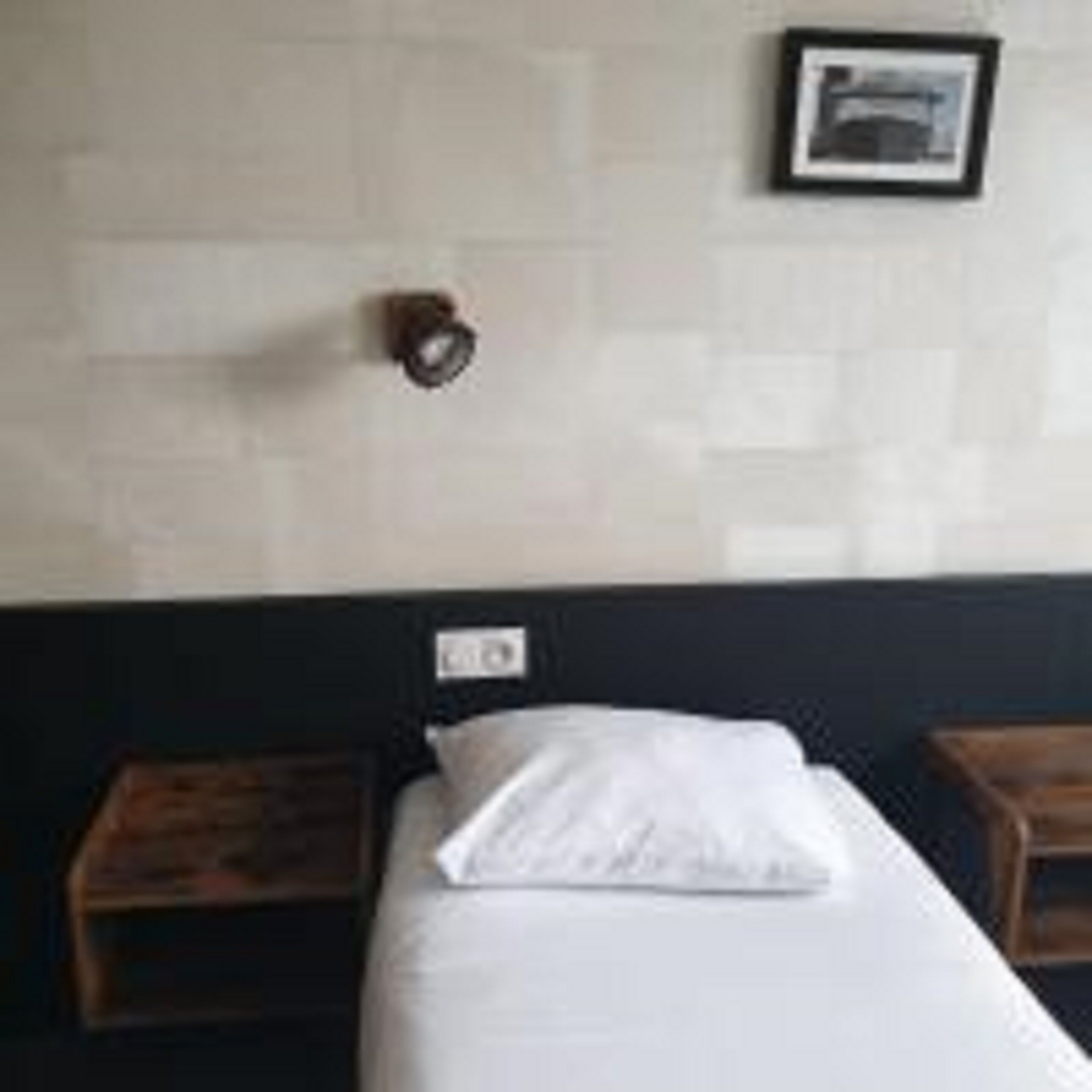 Una cama individual con colchón y almohadas blancas, cabecera de madera, mesita de noche pequeña y lámpara fijada a la pared.