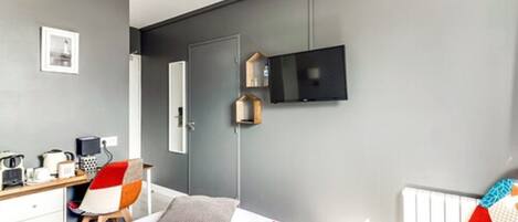 A modern hotel room with a bed, a desk with a computer, a chair, a TV mounted on the wall, and a window.