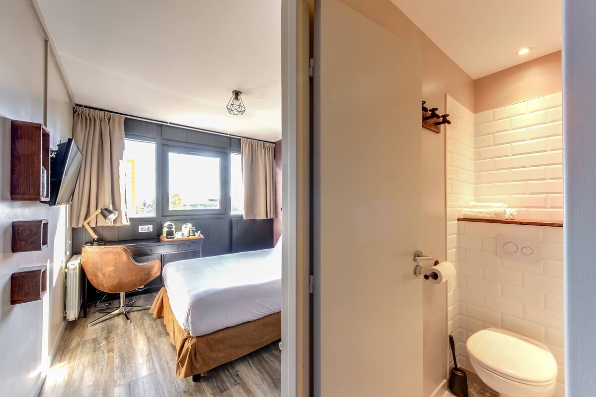 A modern hotel room with a bed, a desk, a chair, a window with curtains, a radiator, and a bathroom with a toilet and sink.