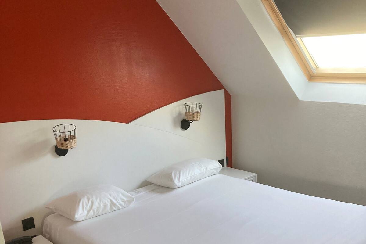 A bedroom with a white bed, a nightstand, a red accent wall, and a skylight.