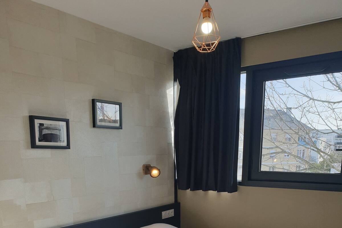 A hotel room with two single beds, a nightstand, a wall-mounted lamp, and a window with curtains.