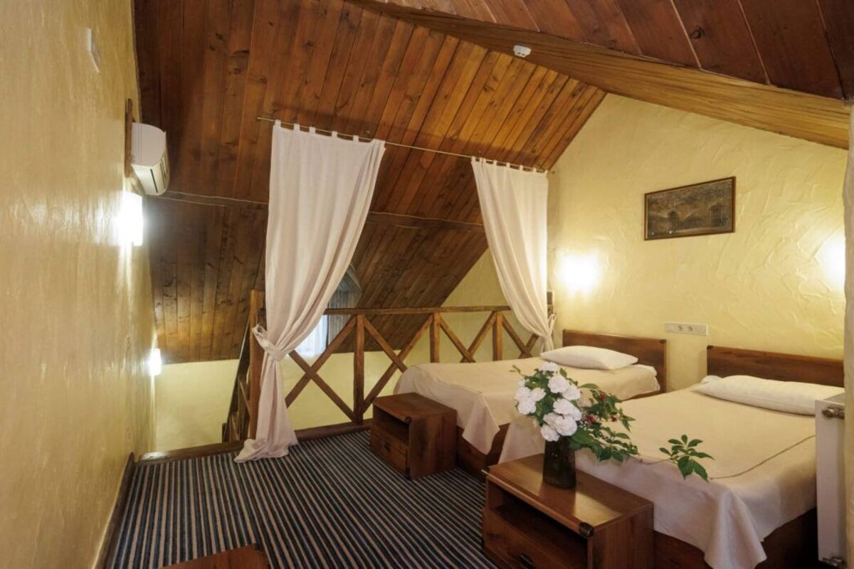 A cozy room with two beds, wooden beams, a small table with a vase, and a window with curtains.