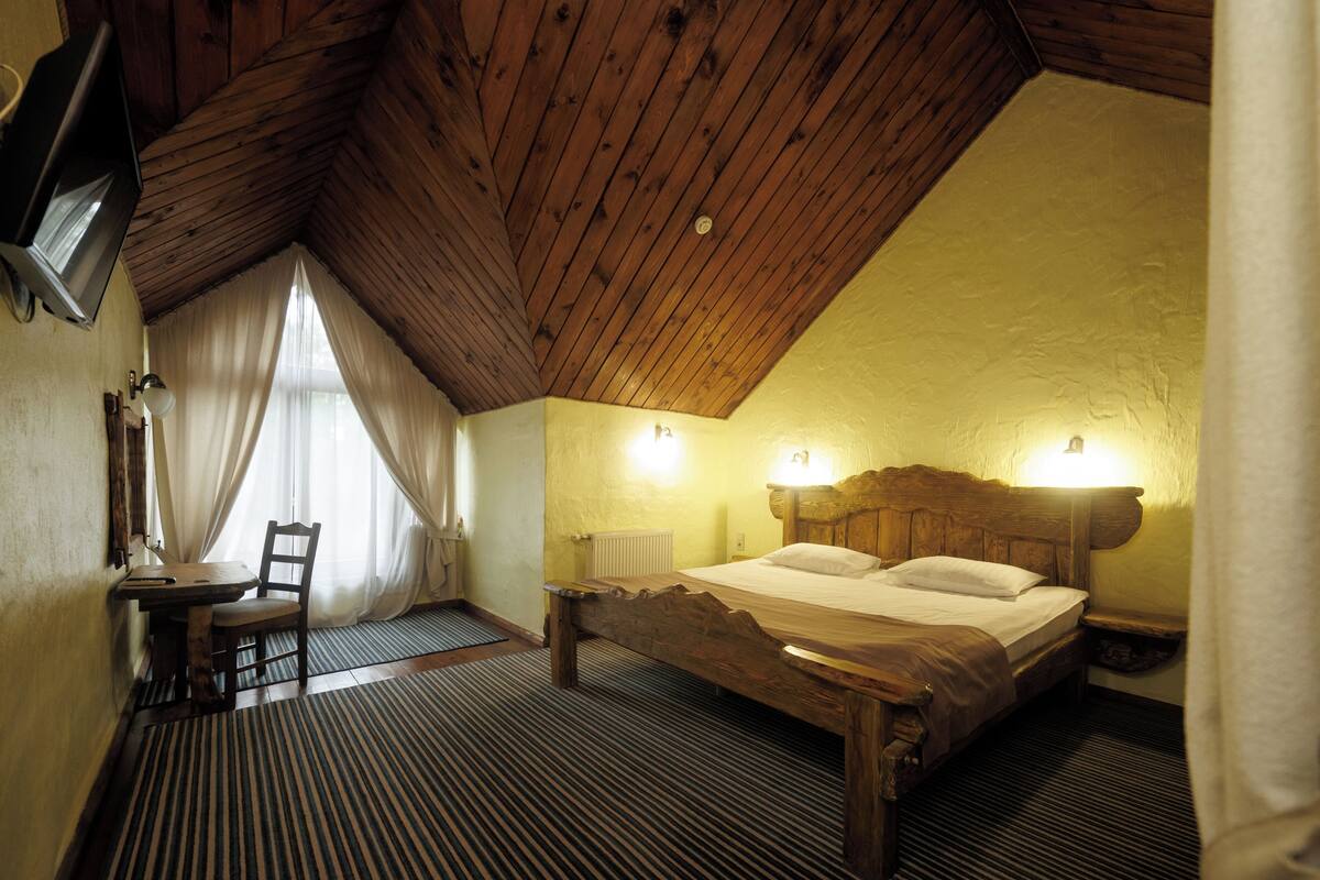 A bedroom with a wooden ceiling, a bed with a wooden headboard, a bedside table, a chair, and a television mounted on the wall.
