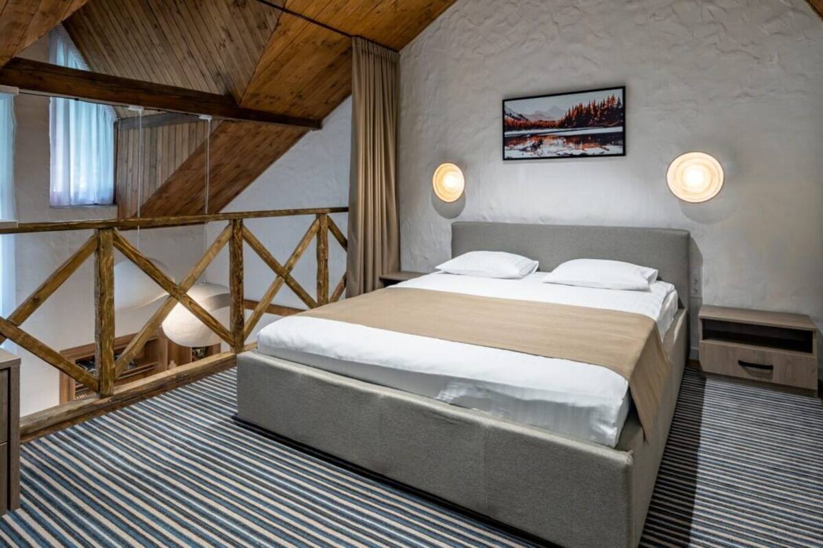A bedroom with a bed, wooden beams, a painting, and two wall sconces.