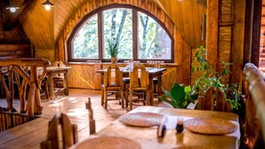 A rustic wooden dining area with a large arched window, wooden tables and chairs, and a central plant. - Park Hotel Drevny Grad (Lviv)
