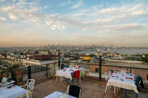 Een terras op het dak met tafels gedekt voor diners, met uitzicht op een stadsbeeld met gebouwen en een waterpartij.