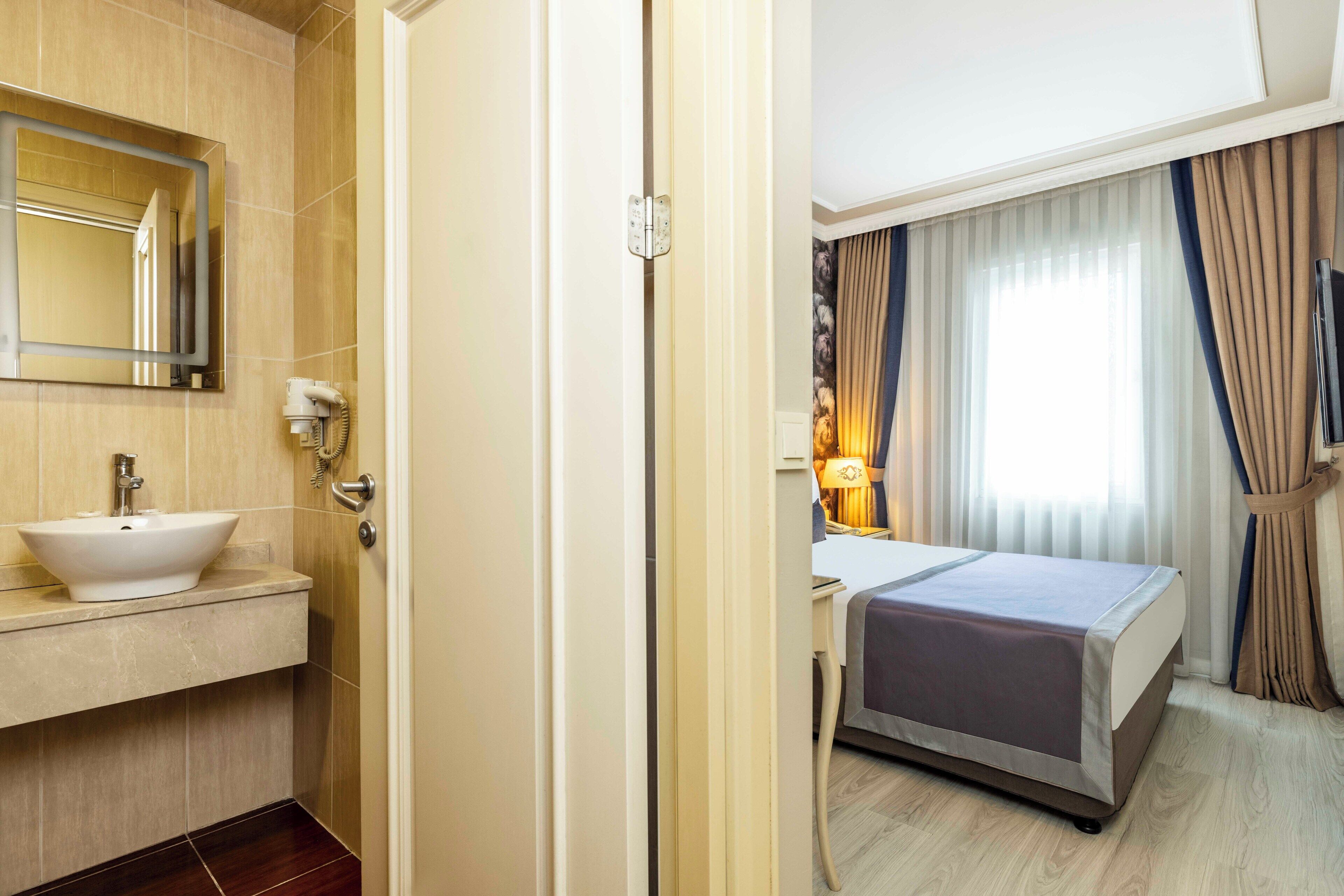 Habitación de hotel con baño que incluye un lavamanos, espejo y inodoro. La habitación tiene una cama, mesita de noche, lámpara y cortinas.