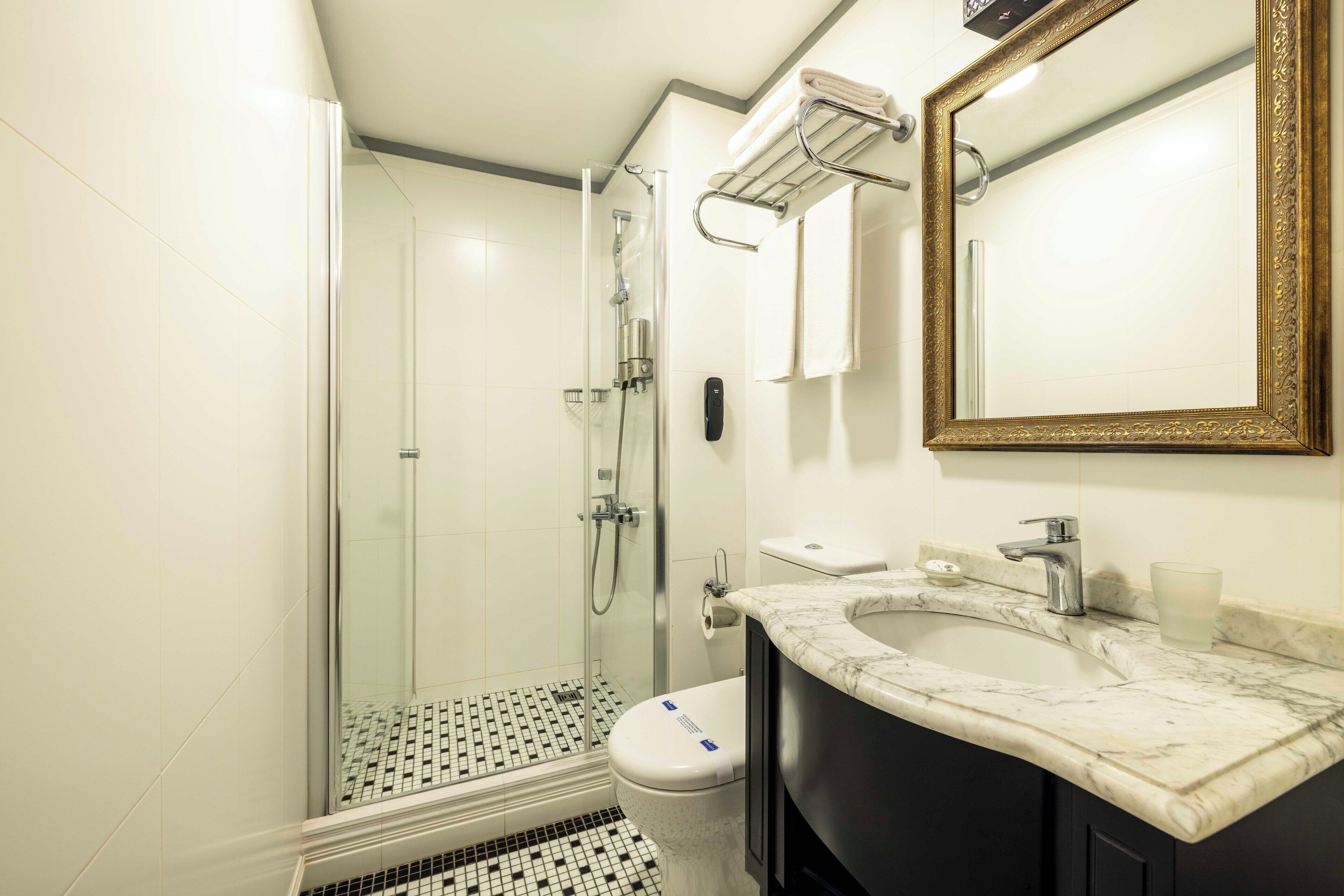 Un baño moderno con ducha de vidrio, lavamanos de mármol y un espejo grande con marco dorado.