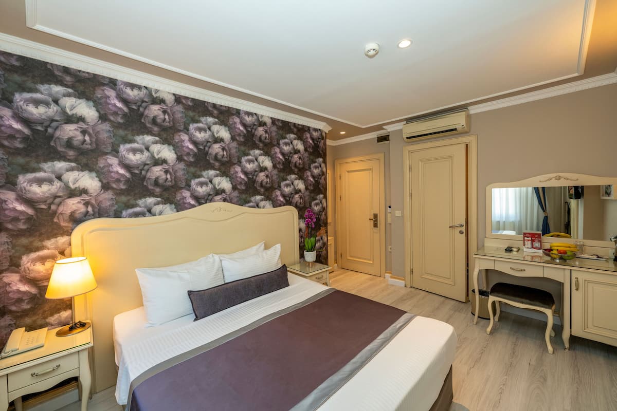 A hotel room with a large bed, a nightstand with a lamp, a dresser, a mirror, and a floral wallpaper.