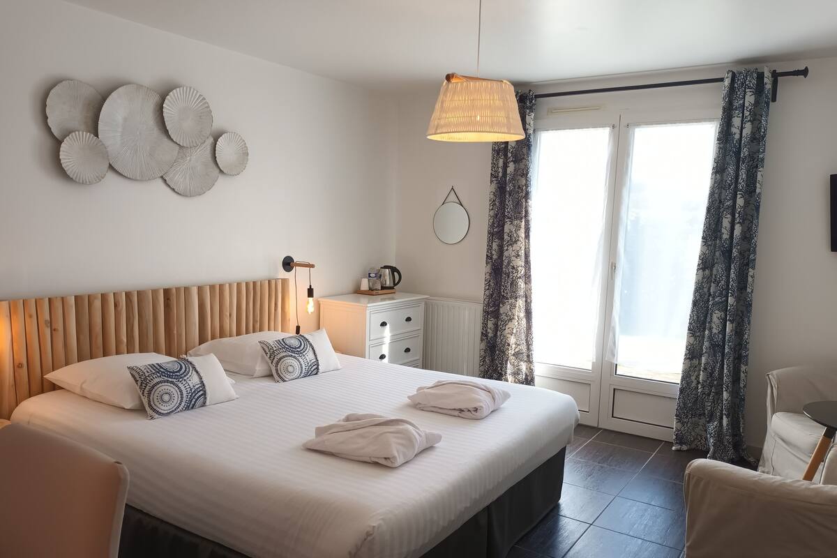 A neatly made bed with pillows, a bedside table with a kettle, a wall-mounted lamp, and a view of the outdoors through a window with curtains.