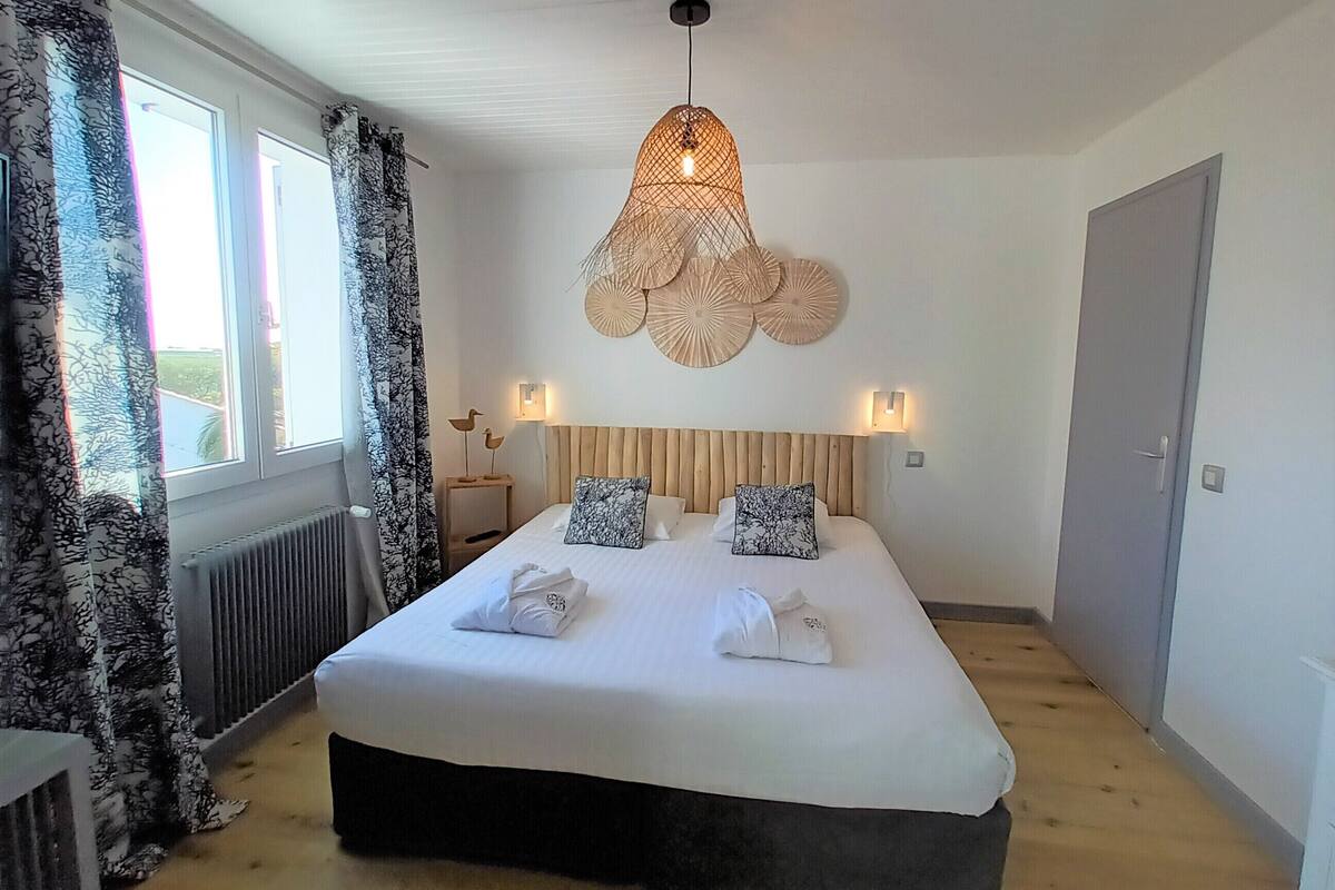 A bedroom with a large bed, a hanging light fixture, and a view of the outdoors through the window.