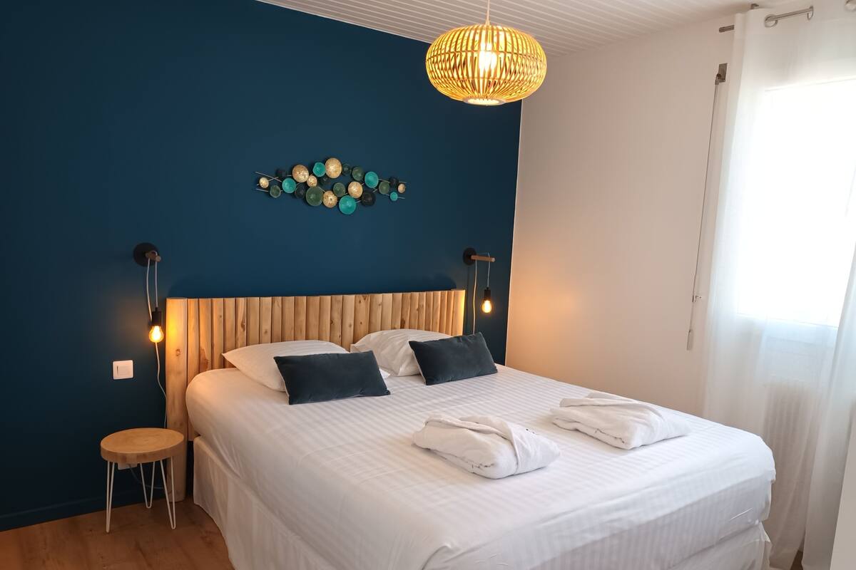 A bedroom with a large bed, two bedside tables, a wall-mounted light fixture, and a window with curtains.