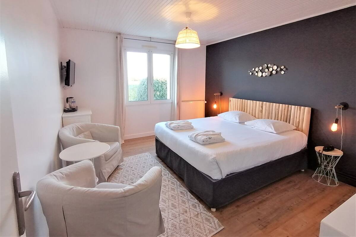 A modern hotel room with a large bed, a seating area with two chairs, a small round table, and a large window.