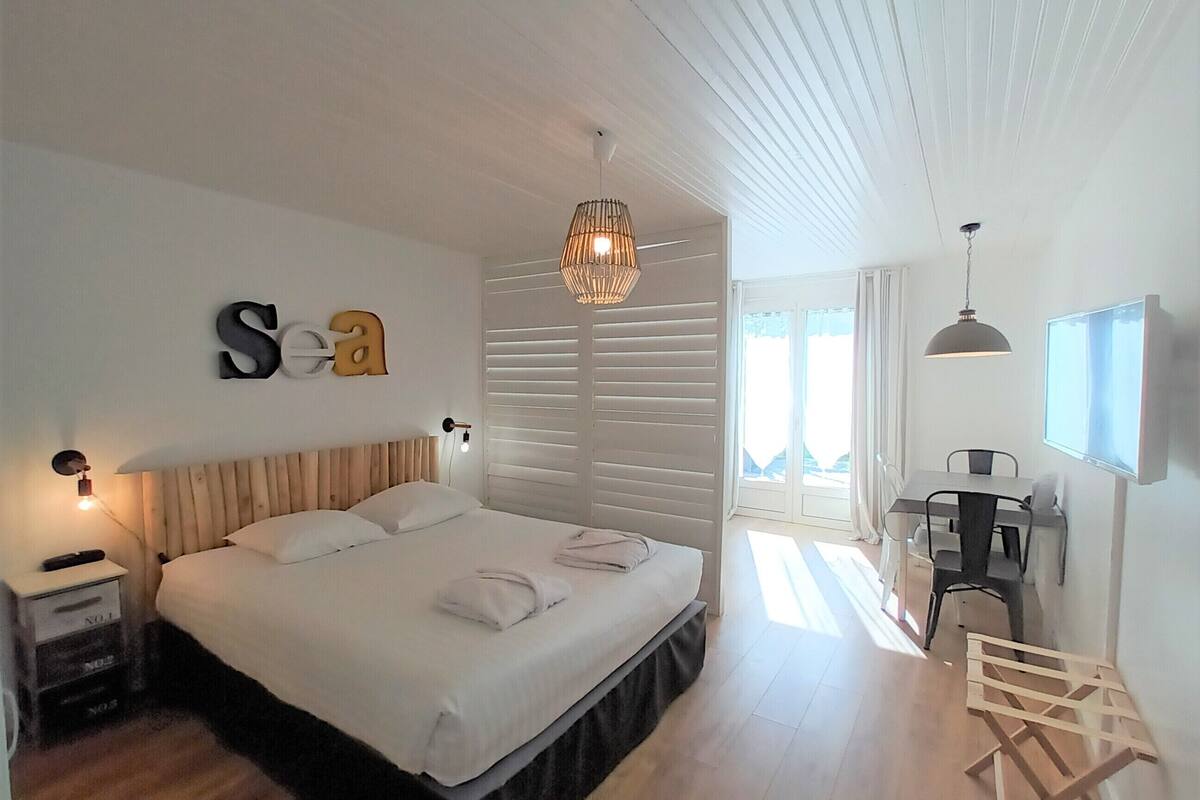 A bedroom with a bed, a nightstand, a desk, and a chair. There is a window with blinds, a ceiling fan, and a wall-mounted TV.