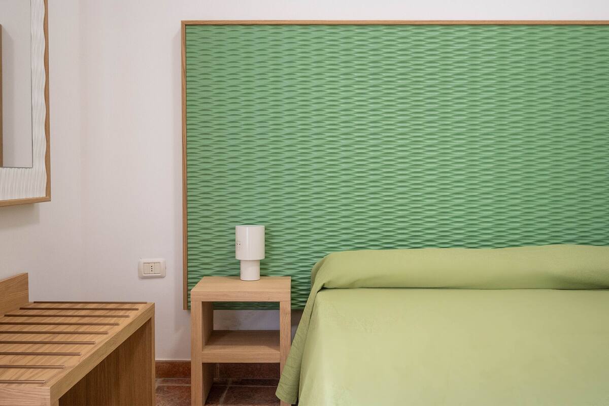 Una habitación con cama, mesita de noche de madera, lámpara y pared verde con textura.