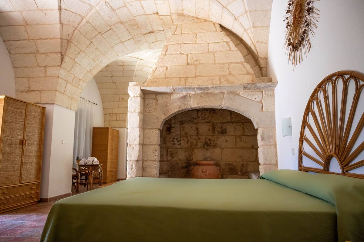 Una habitación con techo abovedado de piedra, cama con colcha verde, cabecero de mimbre y armario de madera.