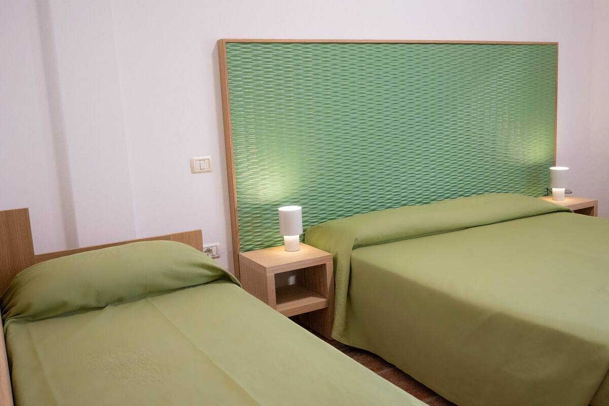Dos camas individuales con ropa de cama verde, cabeceros de madera, mesitas de noche con lámparas y una pared verde con textura.