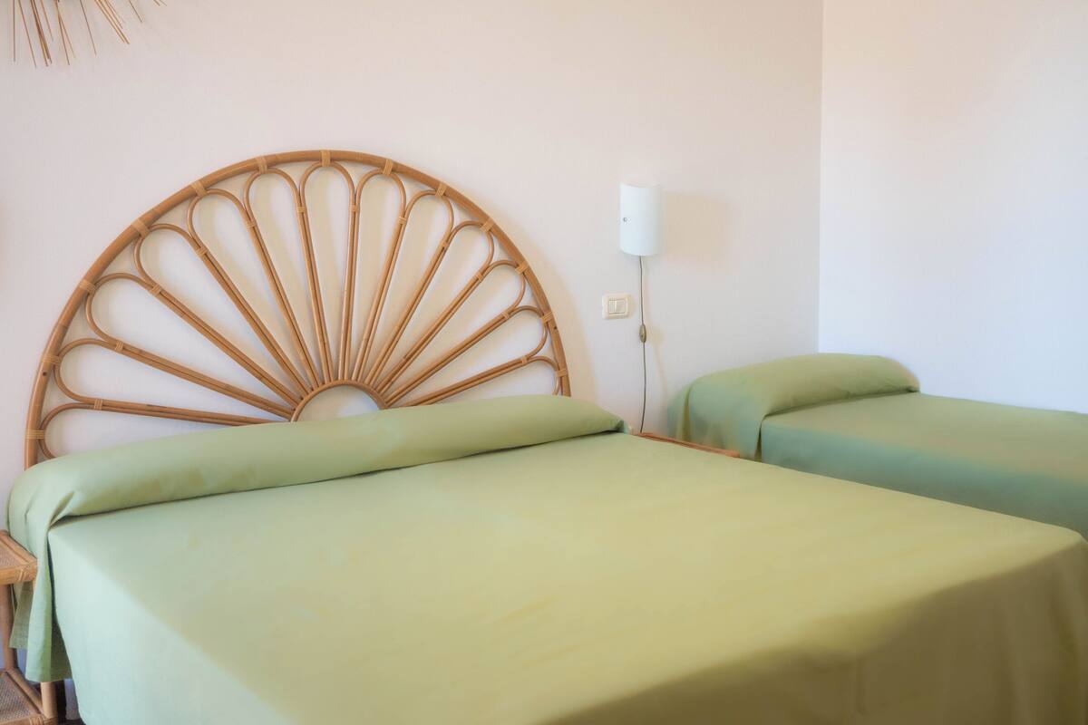 Una habitación con un cabecero circular grande y tejido, dos camas individuales con ropa de cama verde, una mesita de noche y una lámpara fijada a la pared.