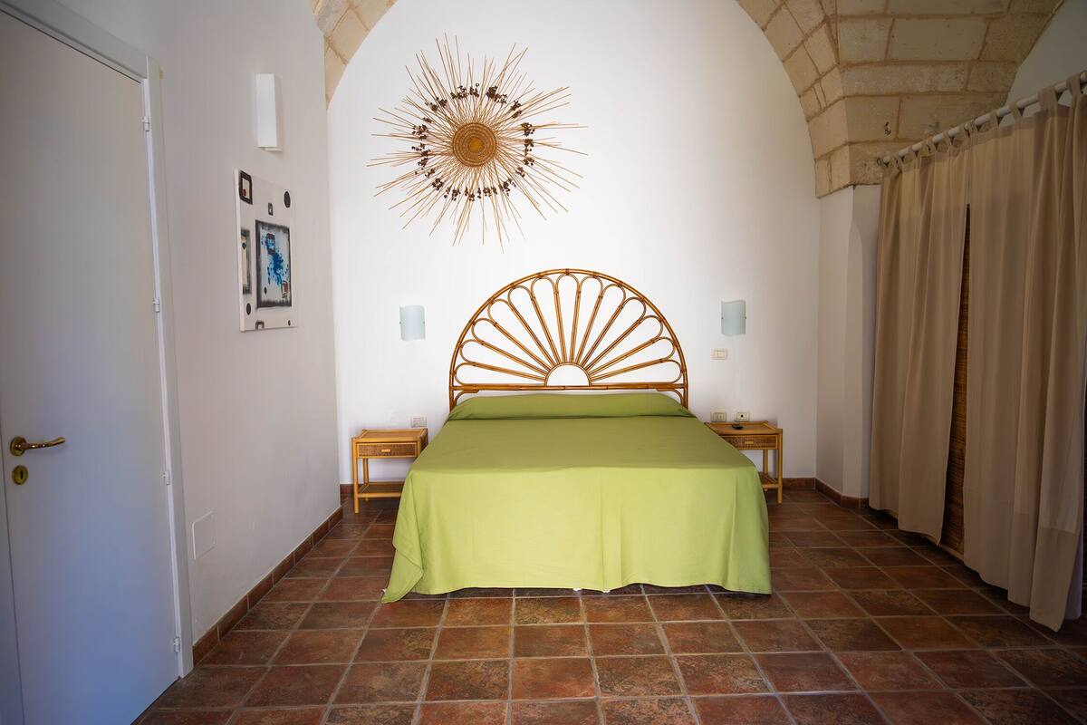 Un dormitorio con una cama, un adorno decorativo con forma de sol en la pared, una puerta y suelo de baldosas.