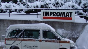 Una furgoneta cubierta de nieve con la marca "Domovina Hotel", estacionada frente a un cartel que dice "IMPROMAT".