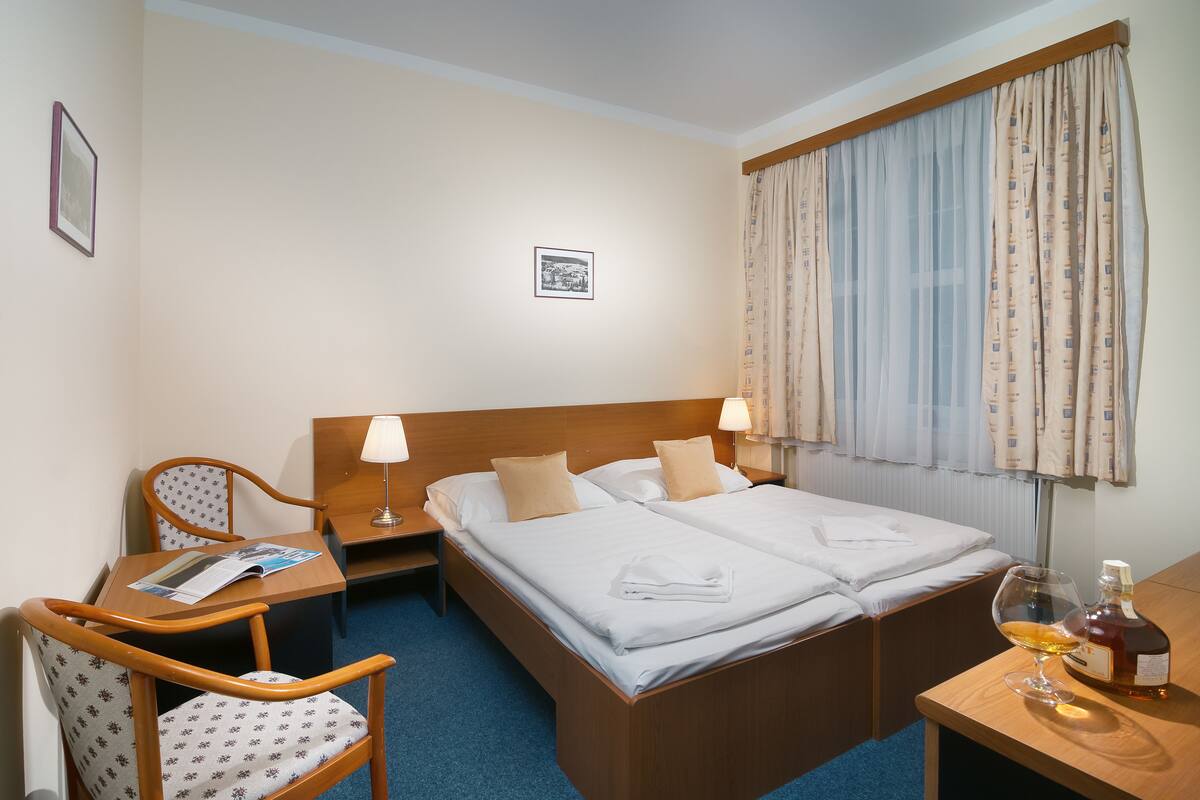 Habitación de hotel con cama doble, dos mesitas de noche, una silla, una pequeña mesa con lámpara y un cuadro en la pared.