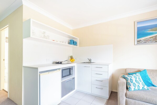 A compact kitchen with white cabinets, a microwave, and a sink. There is a small countertop with a frying pan and a bowl of fruit. A sofa with a patterned cushion is visible in the foreground.
