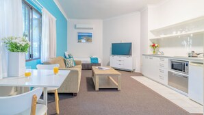 A modern, well-lit living room with a sofa, coffee table, and TV. A kitchen area with a microwave and sink is visible. - Byron BaySide Central Studio Apartments (Byron Bay)