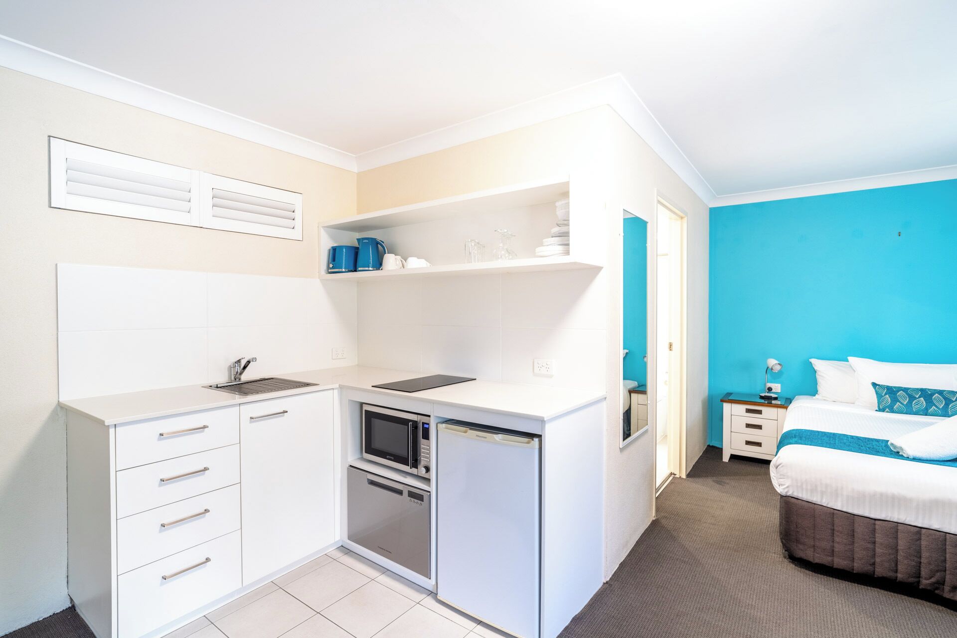 A compact hotel room with a kitchenette, a bed with blue bedding, and a bathroom with a shower.