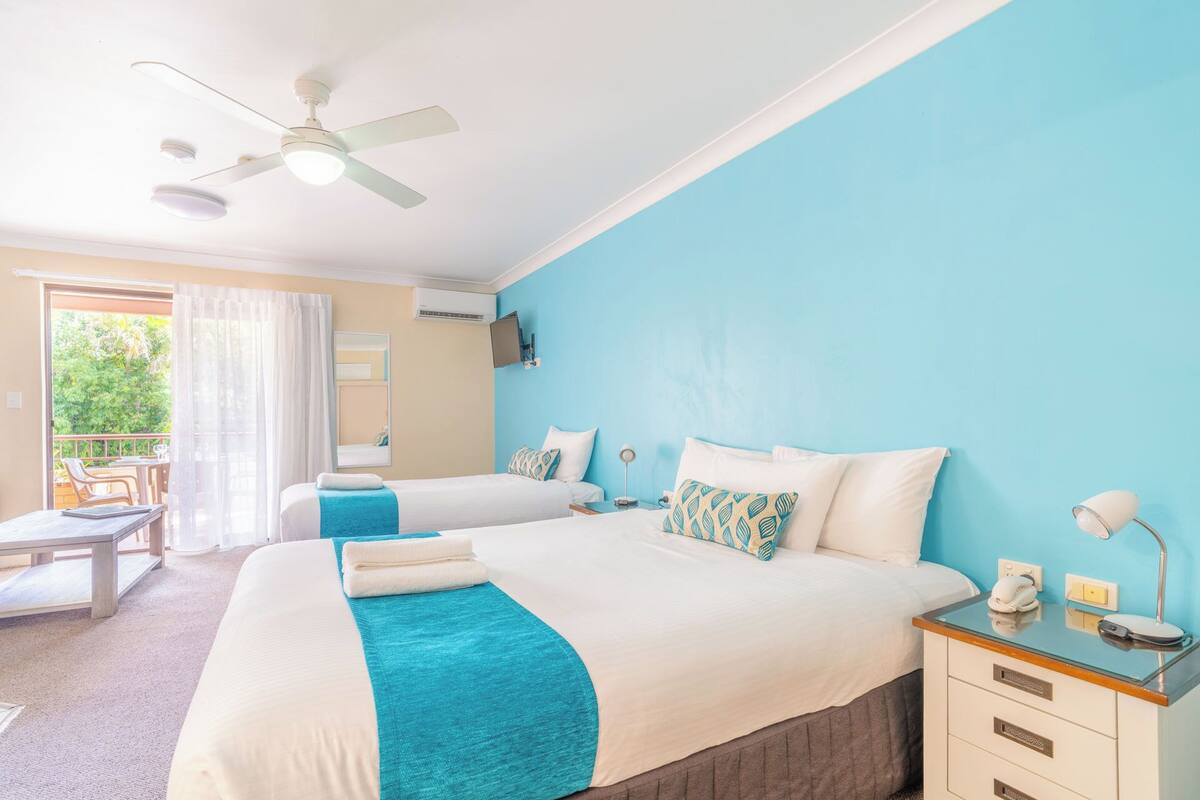 Habitación de hotel con dos camas, ventilador de techo, televisión y balcón con mesa y sillas.