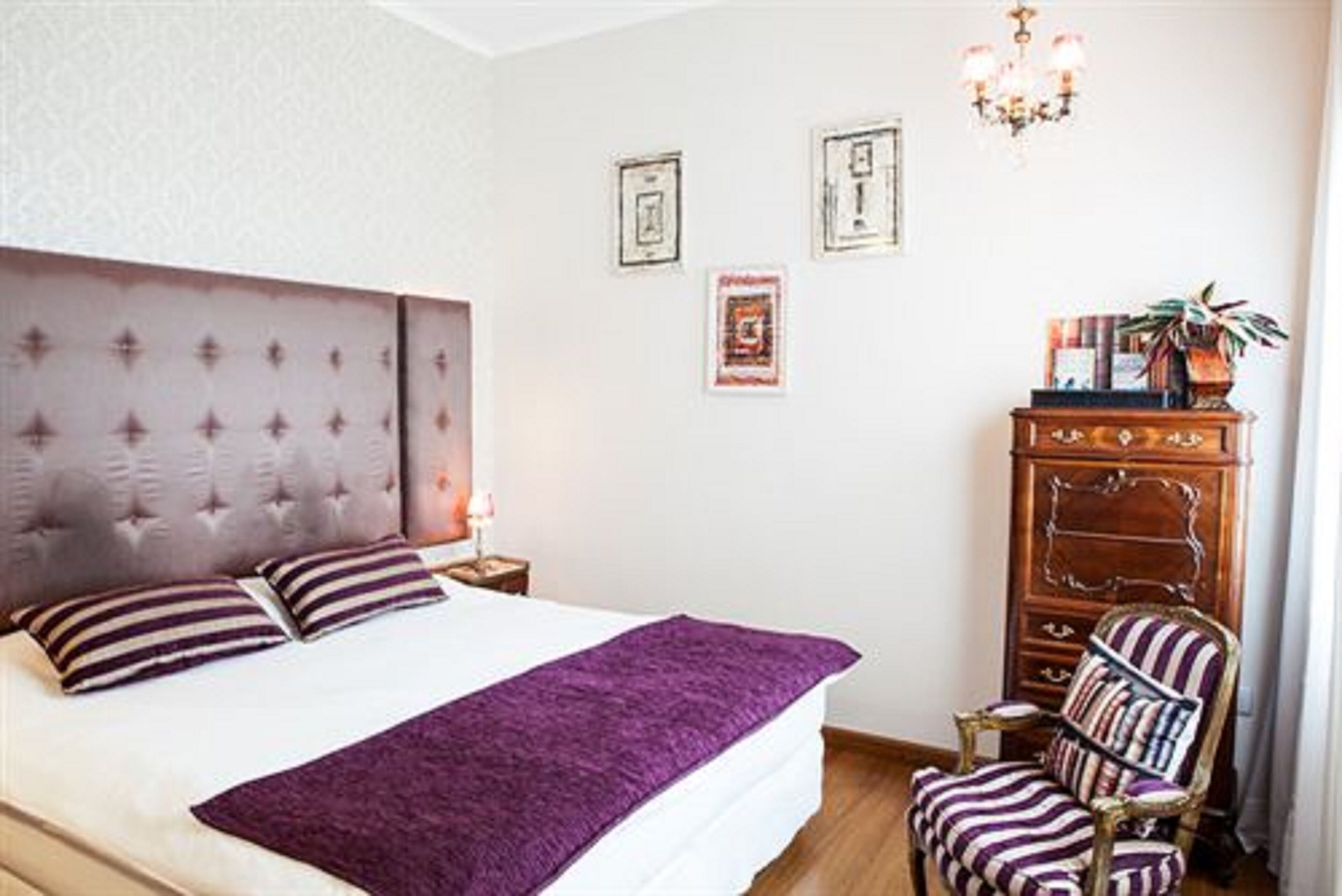 Un dormitorio con cabecera capitonada, cama con almohadas a rayas, manta morada y cómoda de madera, además de un sillón a rayas.