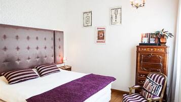 Un dormitorio con cabecera capitonada, cama con almohadas a rayas, manta morada y cómoda de madera, además de un sillón a rayas.
