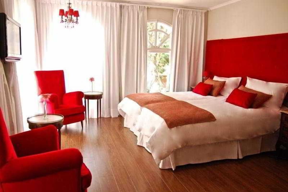 A hotel room with a large bed, red chairs, a small table, and a window with curtains.