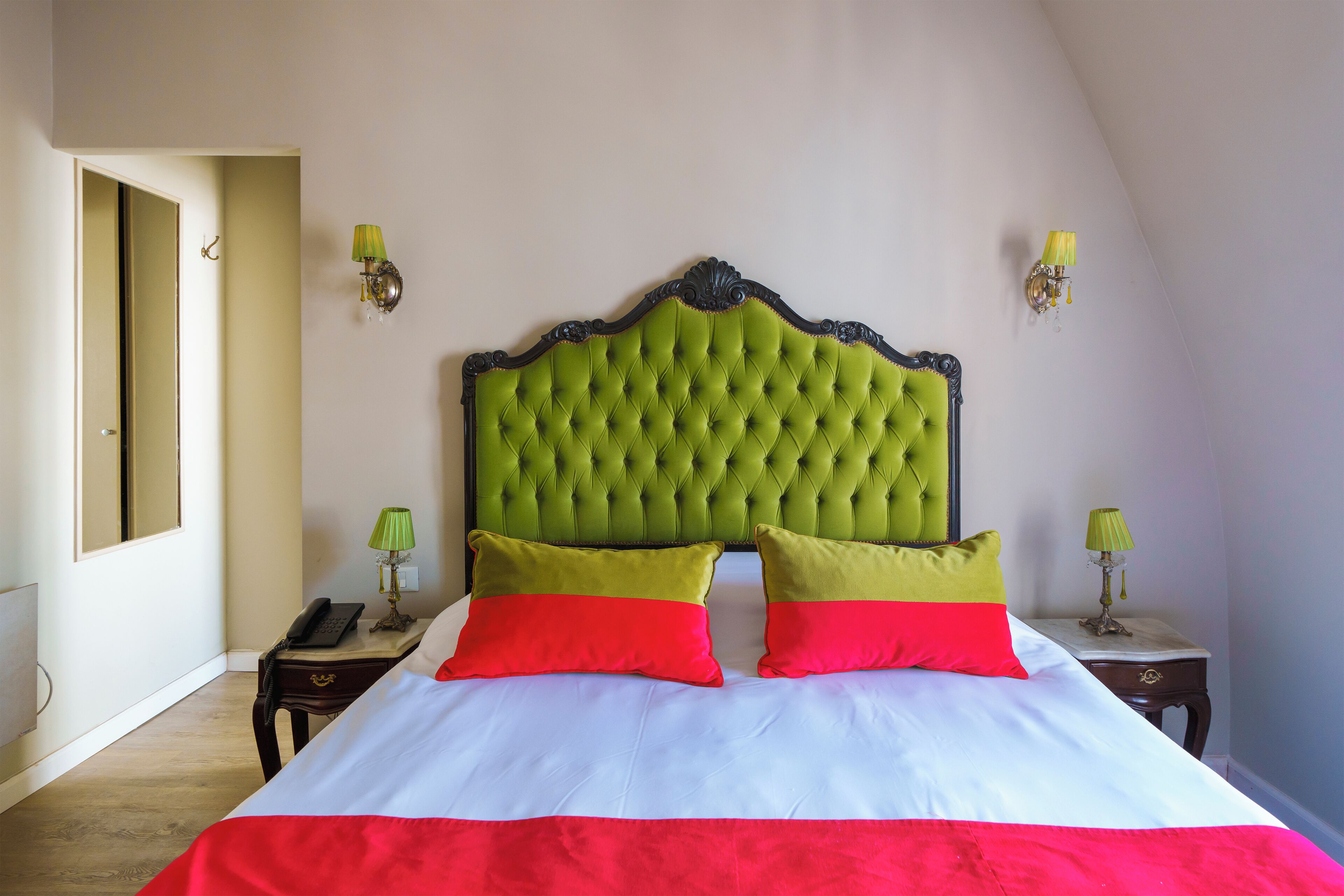 A bedroom with a green tufted headboard, two bedside tables with lamps, and a bed with red and white bedding.