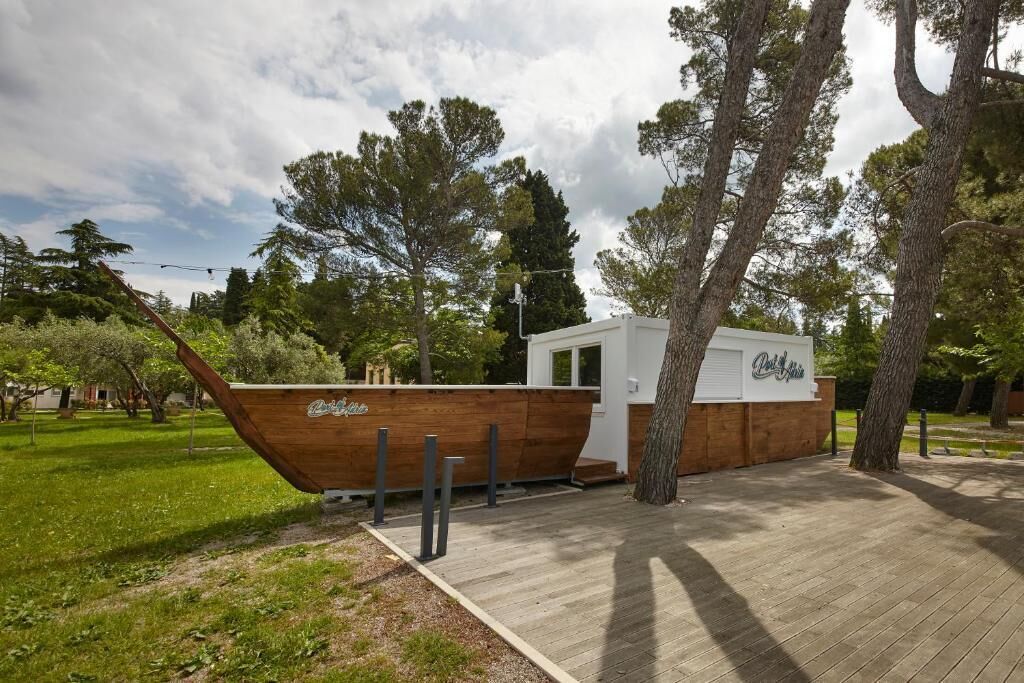 A wooden boat-shaped building with a small window and a door, surrounded by trees and a grassy area.